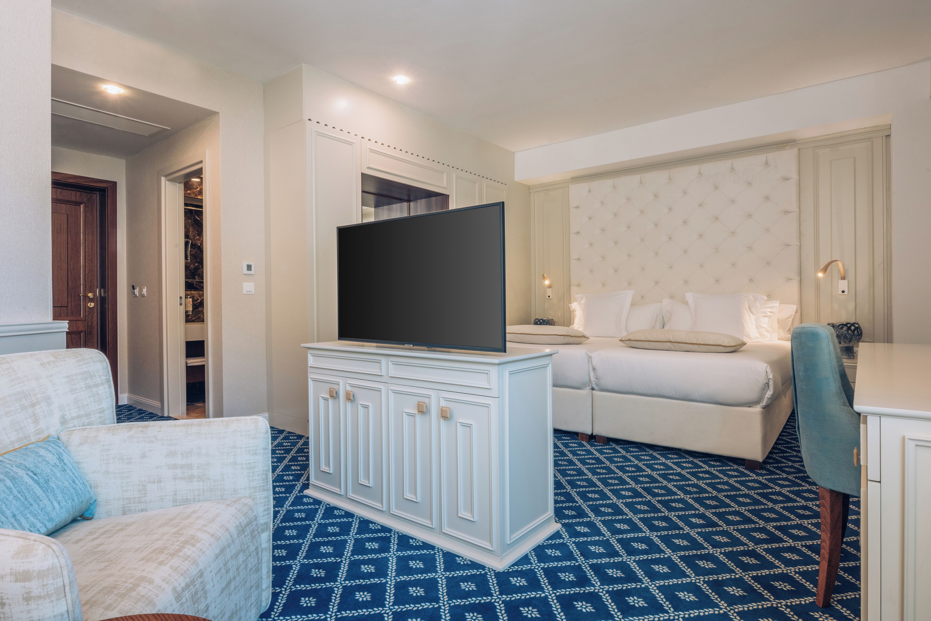 A hotel room with a large flat-screen TV on a cabinet, a bed with white bedding, a blue patterned carpet, and a white armchair.