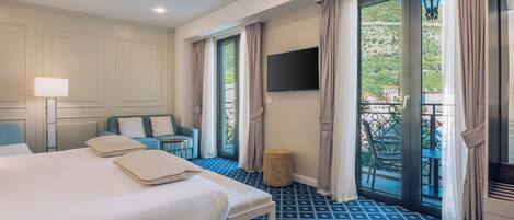 A hotel room with a large bed, a small table, a sofa, a TV mounted on the wall, and a balcony with a view of the outdoors.