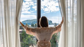 Une personne, les bras tendus, se tient face à un paysage magnifique, visible à travers une fenêtre ornée de rideaux légers.
