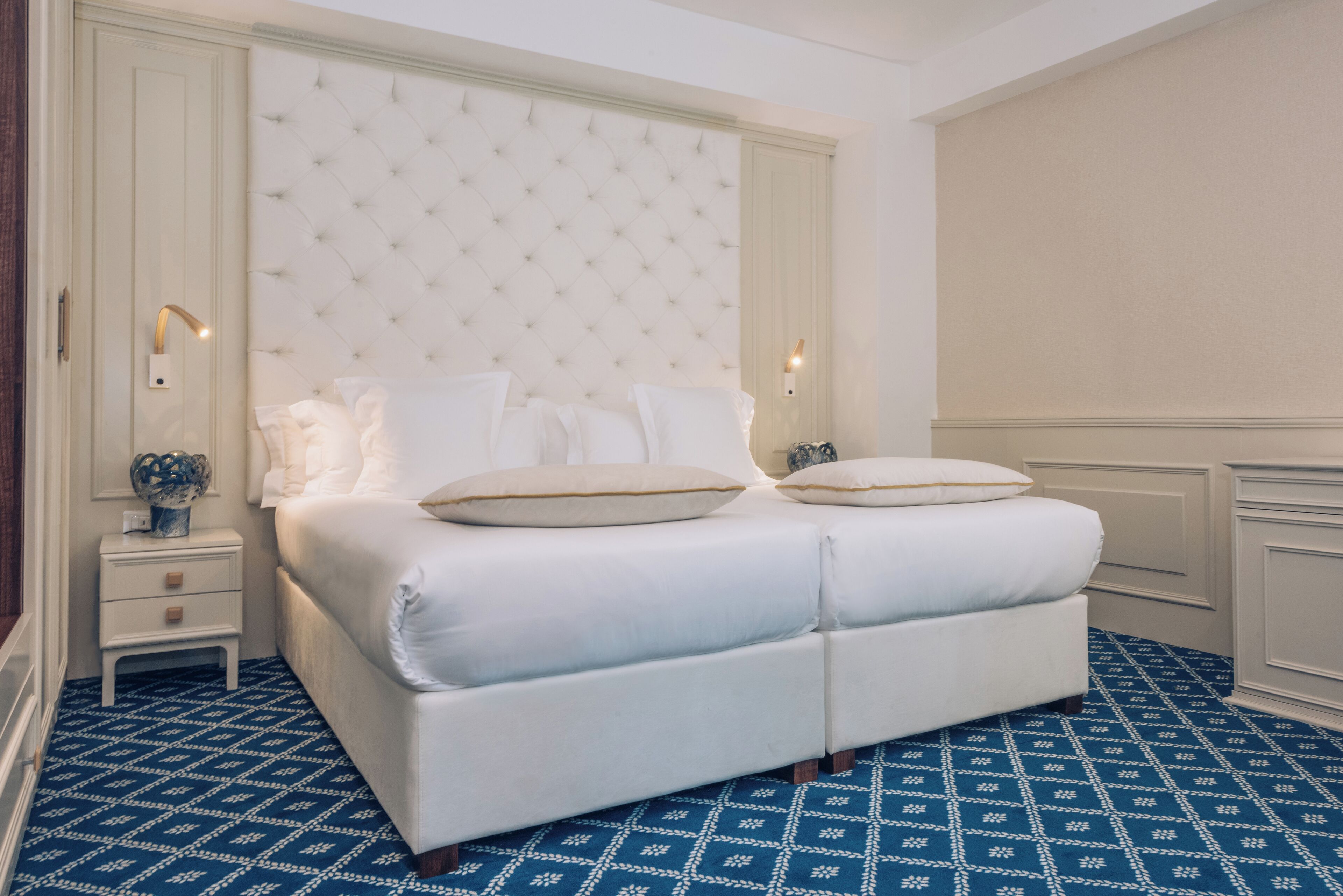 A large, white, tufted headboard bed with two pillows, a nightstand with a lamp, and a patterned blue carpet.