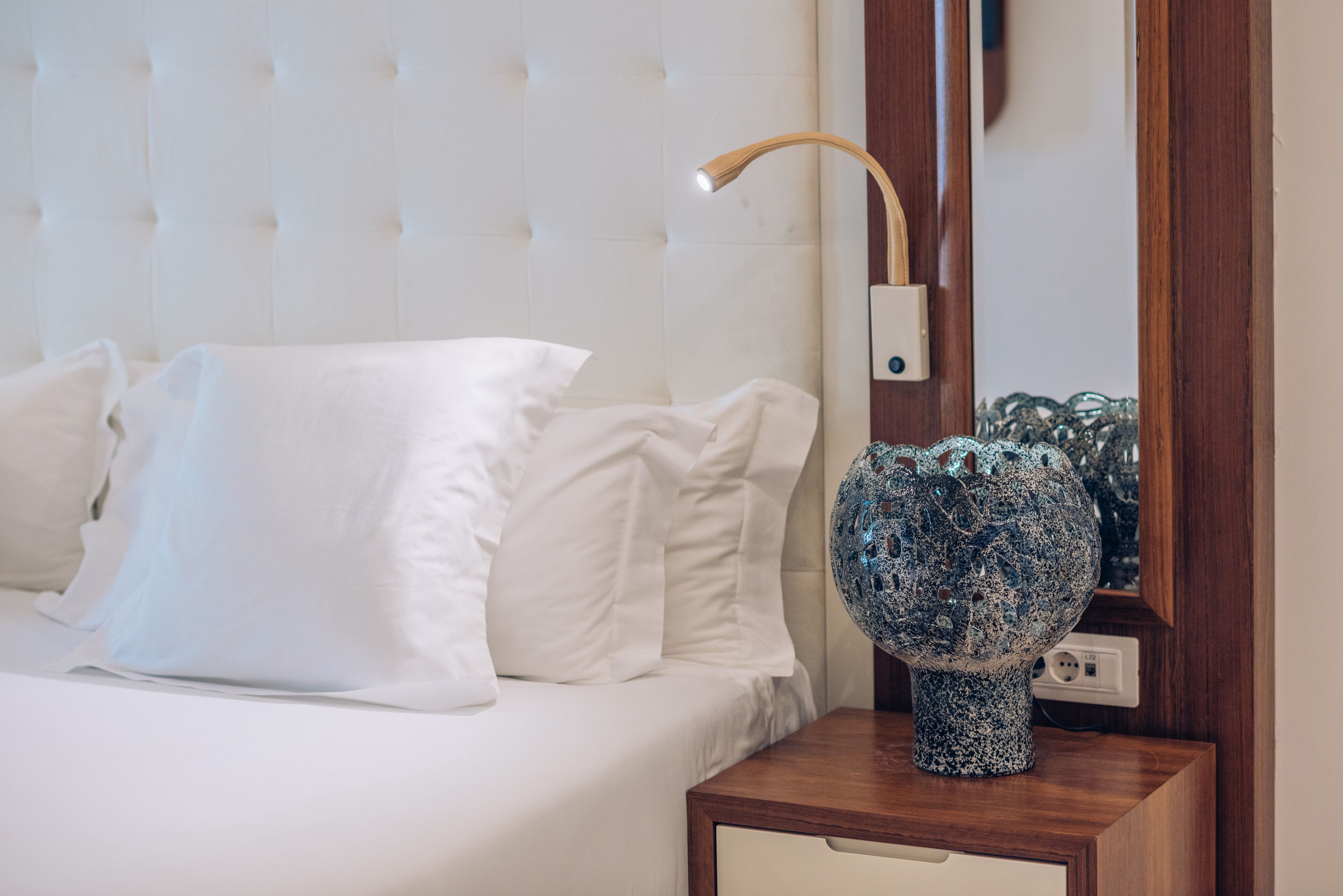 Una cama bien hecha con sábanas blancas, una mesita de noche de madera con un jarrón decorativo y un cabecero moderno.