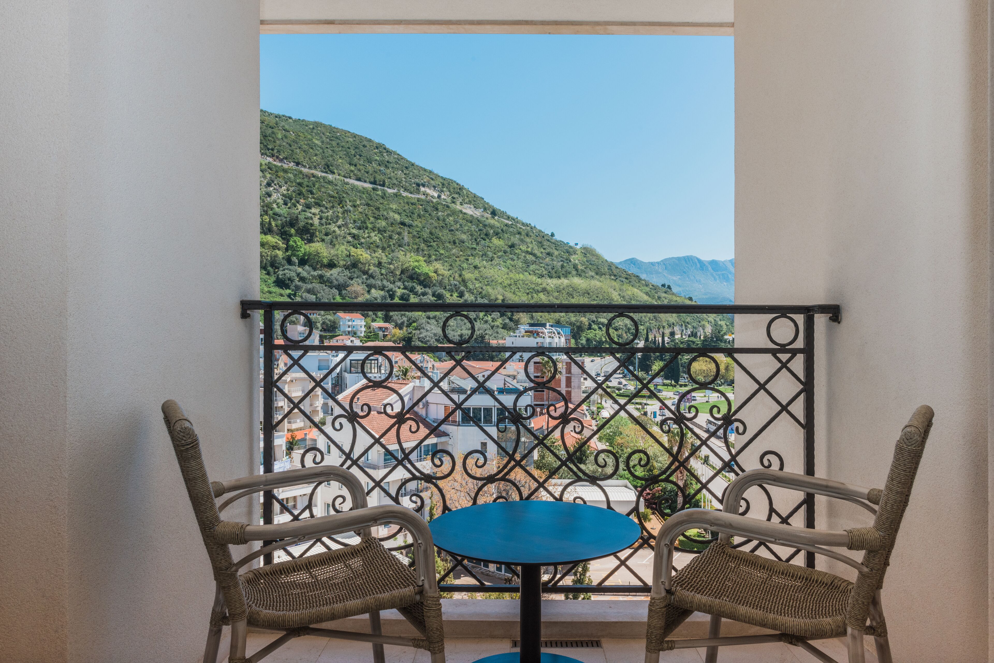 Un balcon agrémenté de fauteuils en osier et d’une petite table, d’où l’on peut admirer un paysage montagneux parsemé de bâtiments.