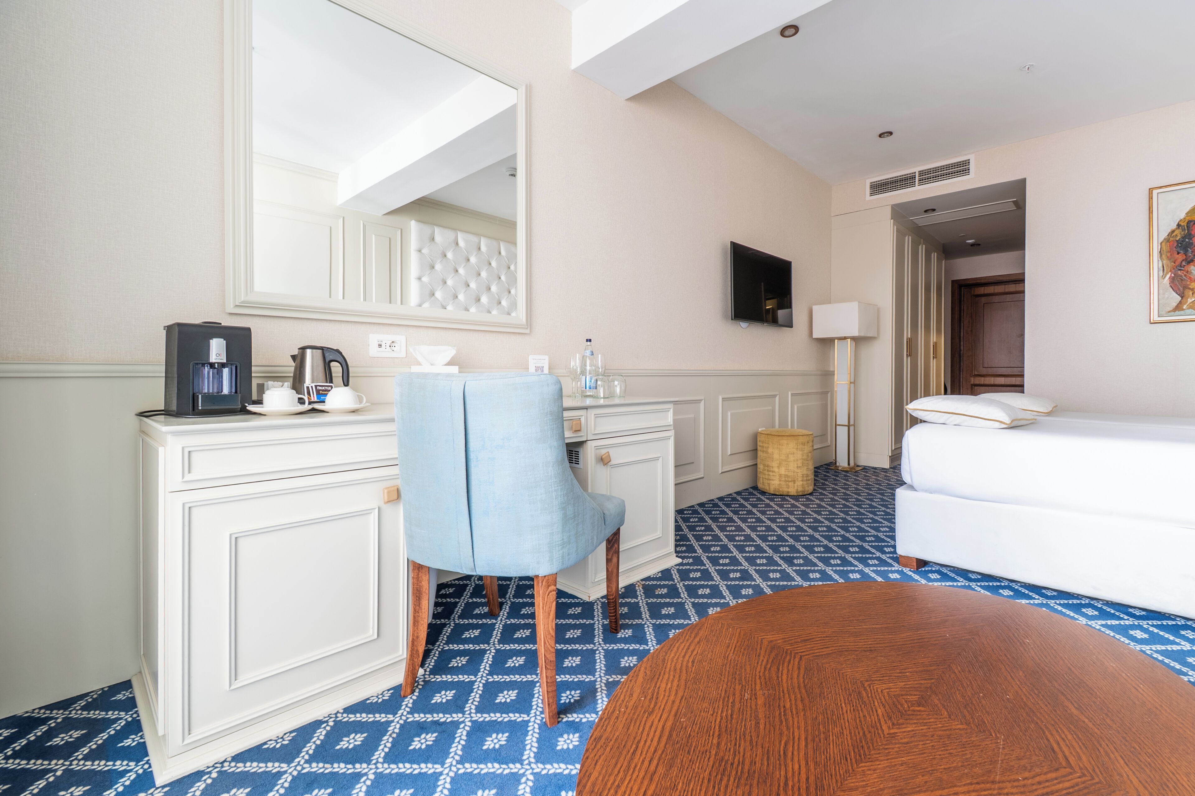 A hotel room with a white dresser, a blue armchair, a white sofa, a wooden coffee table, a flat-screen TV, and a patterned floor.