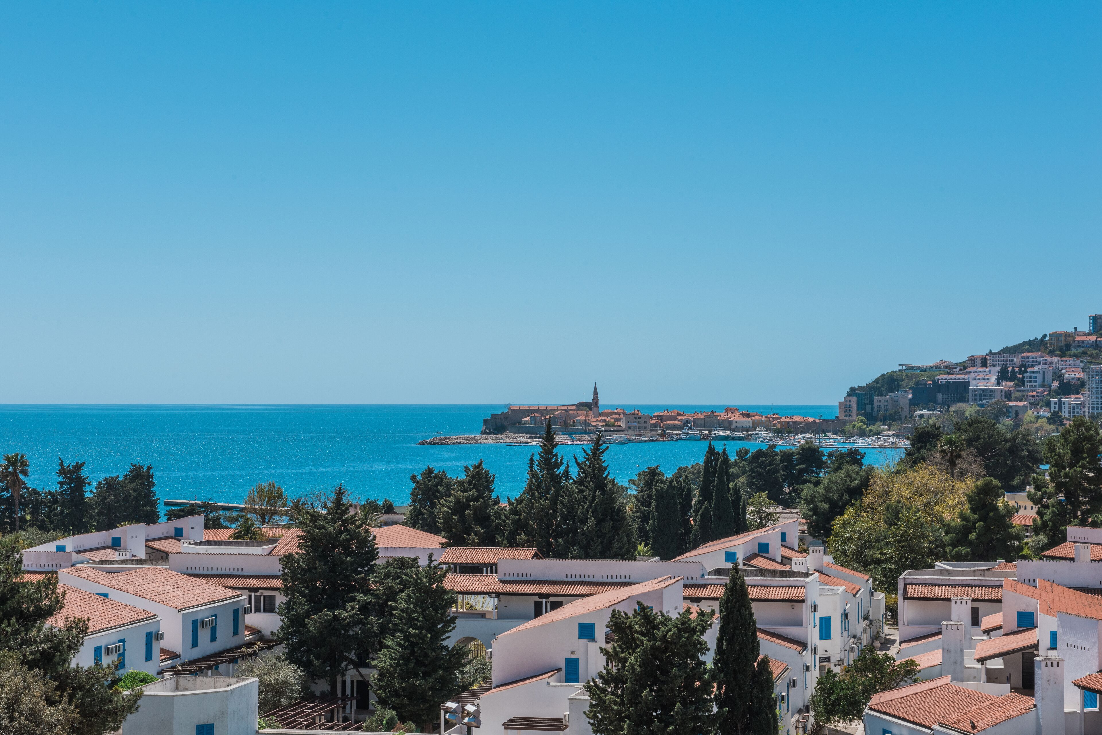 Un pueblo costero con edificios blancos y tejados rojos, un cielo azul despejado, y vista al mar y una isla distante.