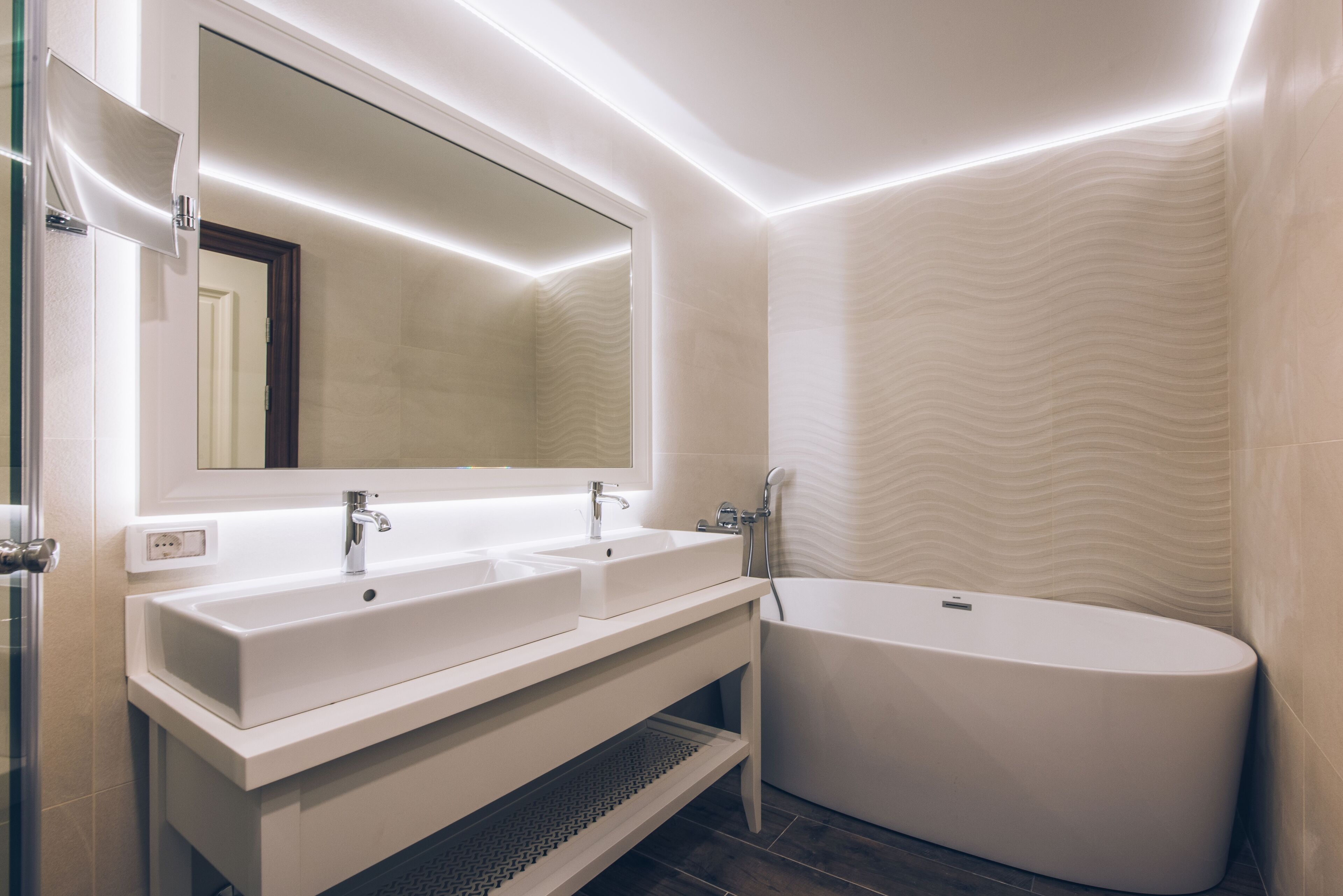 A modern bathroom with a large mirror, two sinks, a bathtub, and a textured wall.