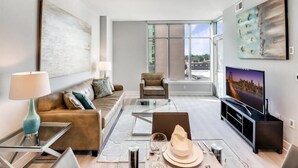 A modern living room with a sofa, a glass coffee table, a dining table set for two, and a television. - Easy Travel to DC | Full Kitchen, Pool & Spa | Downtown Bethesda (Bethesda)