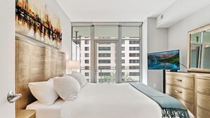 Moderni hotellihuone, jossa on suuri sänky, televisio, puinen lipasto ja seinällä kaupunkimaisemaa esittävä taulu.