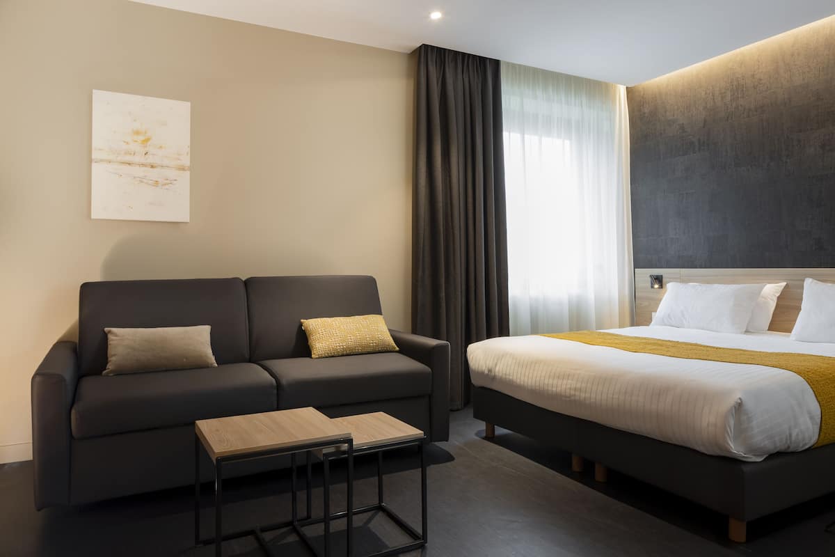Ein modernes Hotelzimmer mit einer dunkelgrauen Couch, einem Holztisch, einem großen Bett mit weißer und gelber Bettdecke und einem großen abstrakten Gemälde an der Wand.