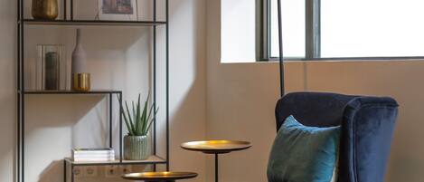 Una habitación moderna con una silla azul oscuro, una mesita redonda pequeña y una alta estantería negra con diversos objetos decorativos.