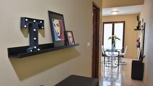 Un couloir avec une étagère noire où sont exposées des photos encadrées et une lettre décorative en forme de T. Une petite table avec un vase et un objet en forme de cube se trouve au premier plan.