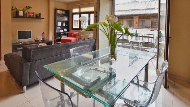 Un salon moderne avec une table à manger en verre, un canapé gris, une bibliothèque et une vue sur le balcon extérieur.
