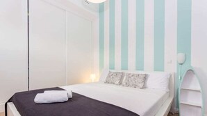 A neatly made bed with a black bedspread and a folded white towel, a white nightstand with a lamp, and a white wardrobe with a teal stripe wall. - Apartment in Vilamoura (Vilamoura)