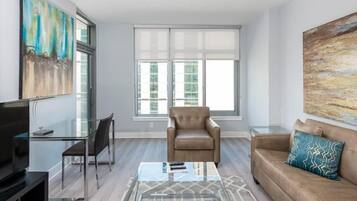 A modern living room with a glass coffee table, leather sofa, and a chair. There is a TV on a stand and artwork on the walls.