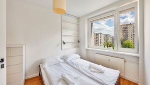 A bedroom with a bed, a window with a view of buildings, a radiator, and a hanging light fixture. - Apartamenty Sun & Snow Gdynia Centrum (Gdynia)