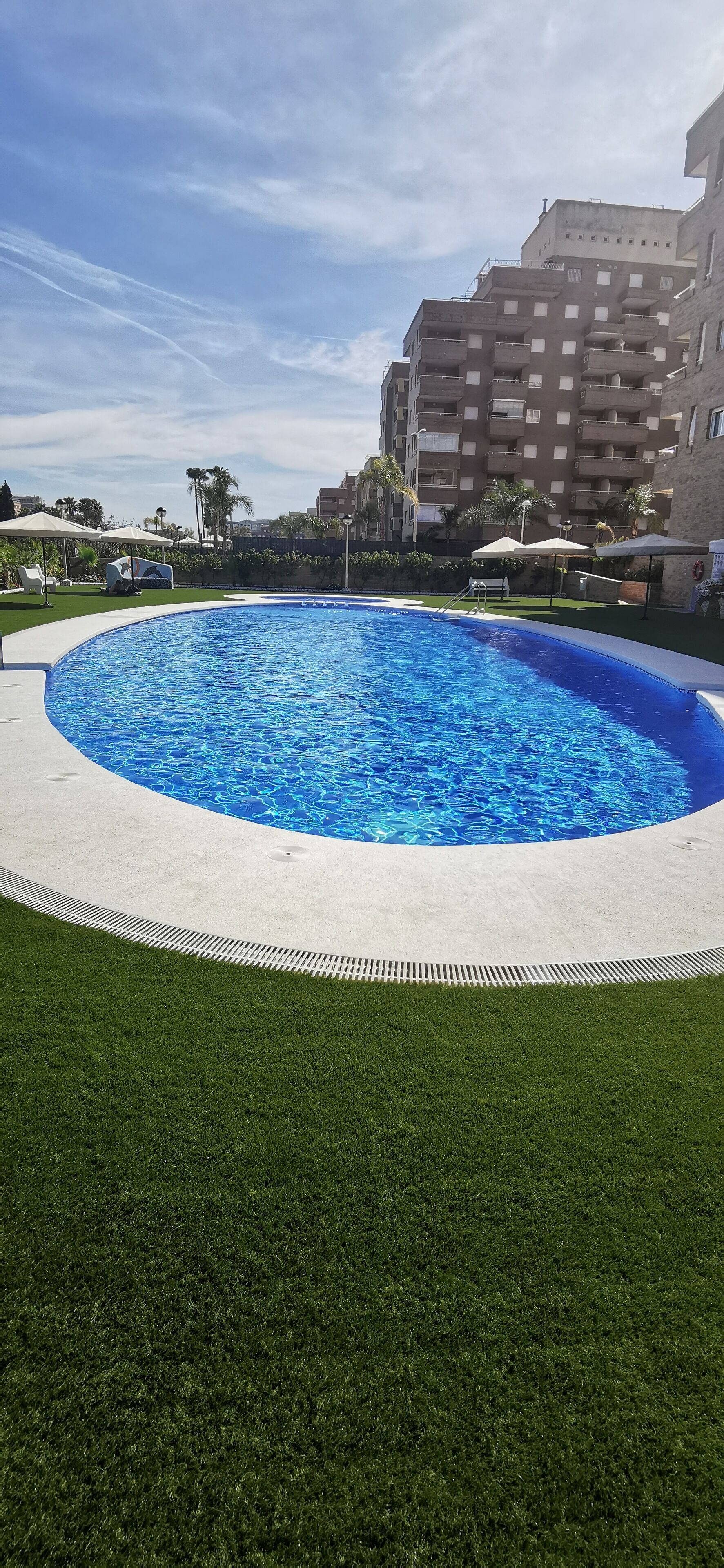 En rund pool med en vit kant, omgiven av grön konstgräsmatta, och med bostadshus i bakgrunden.