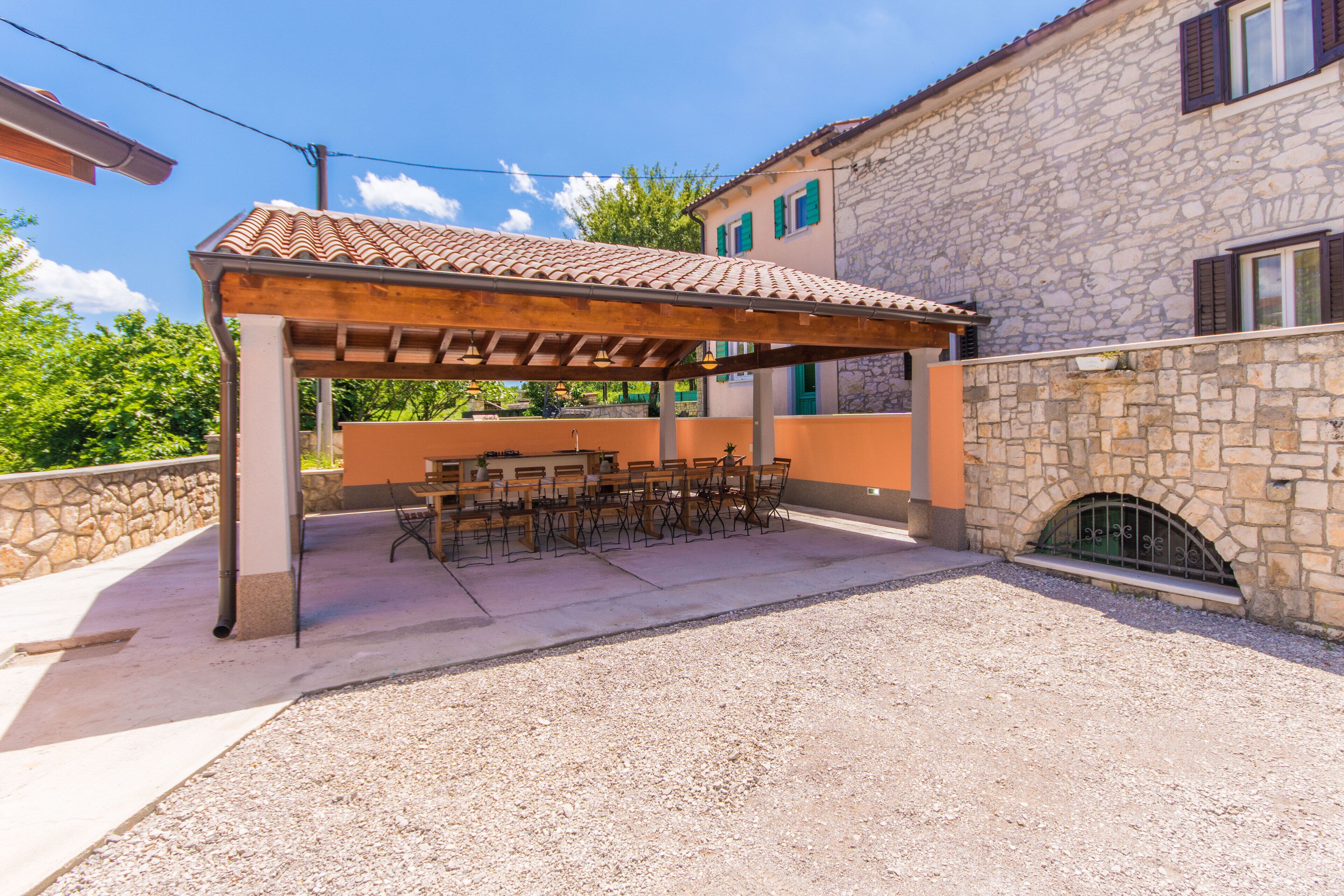 An outdoor patio with a pergola, tables, and chairs, adjacent to a stone building with a tiled roof.