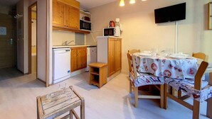 A compact kitchen with wooden cabinets, a microwave, and a refrigerator. There is a dining table with chairs and a small wooden stool. A television is mounted on the wall. - 5th floor, tv, ski locker, 22m², Les Menuires (Les Menuires)