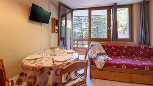 A cozy room with a dining table set for four, a sofa, a TV mounted on the wall, and large windows offering a view of trees. - 5th floor, tv, ski locker, 22m², Les Menuires (Les Menuires)