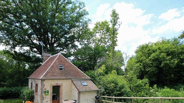 Un petit bâtiment en pierre avec un toit en tuiles, une porte et une fenêtre. On y voit une table et des chaises à l’extérieur, ainsi qu’une clôture en bois à l’arrière-plan.