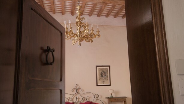 Une chambre avec une porte en bois, un lit avec une literie rouge, un lustre et un tableau encadré au mur.