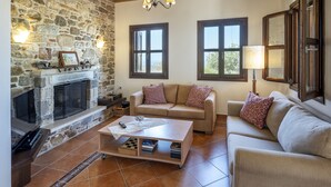 A cozy living room with a stone fireplace, a sofa, a coffee table, and two windows with curtains. - Villa Lagoudi Stone House (Λαγουδι Ζία)