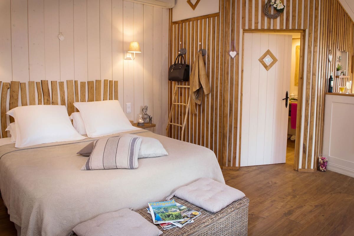 A bedroom with a bed, pillows, a nightstand, a lamp, a wicker basket with magazines, and a wooden door leading to another room.