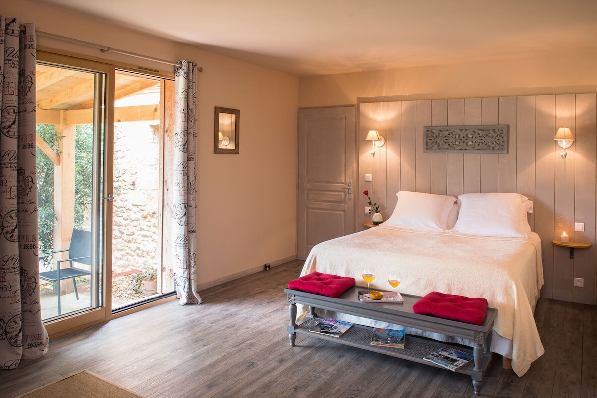 A bedroom with a bed, bedside lamps, a nightstand with a red cushion, a window with curtains, and a door.