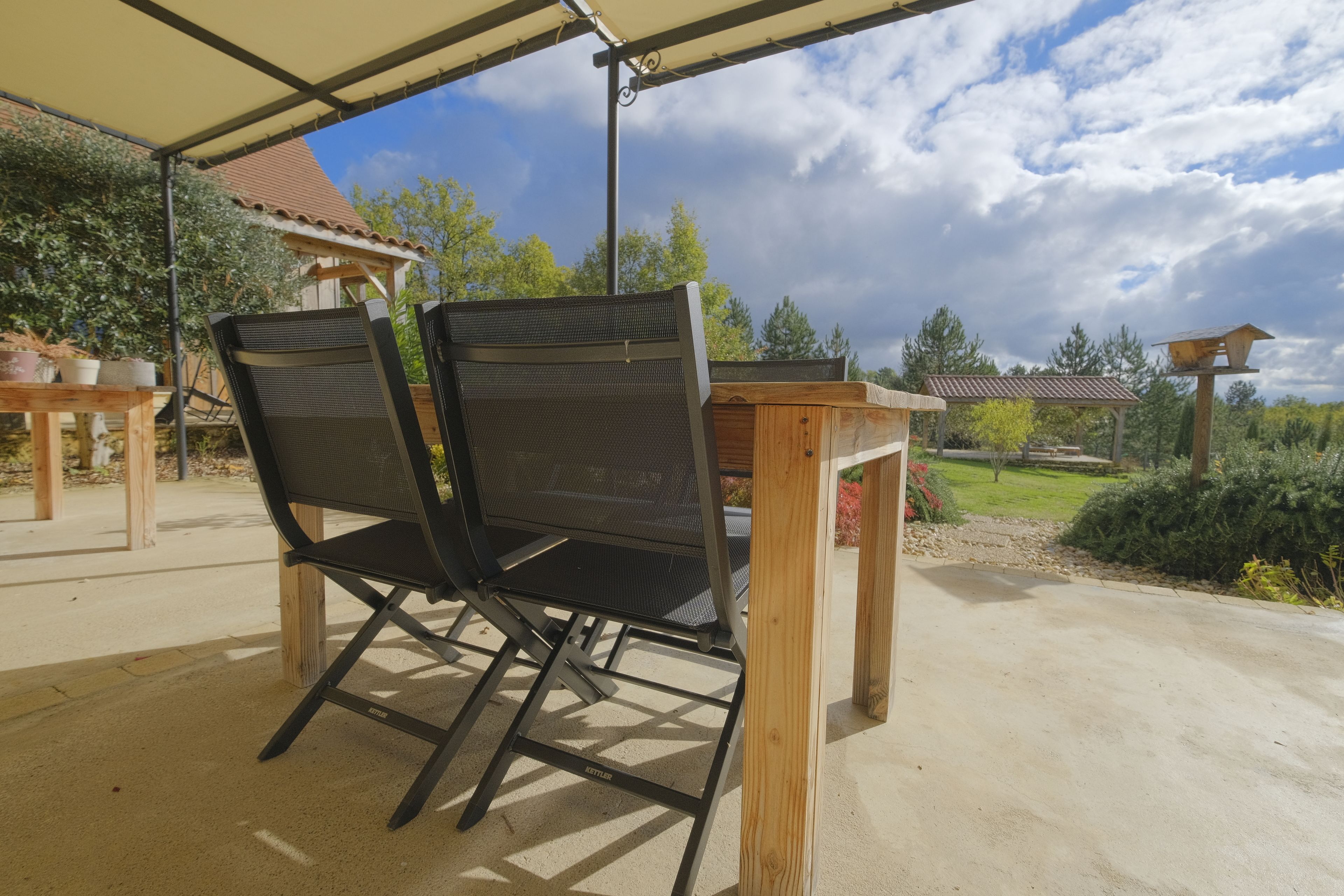 Een buitenpatio met twee zwarte mesh stoelen, een houten tafel en een pergola erboven.