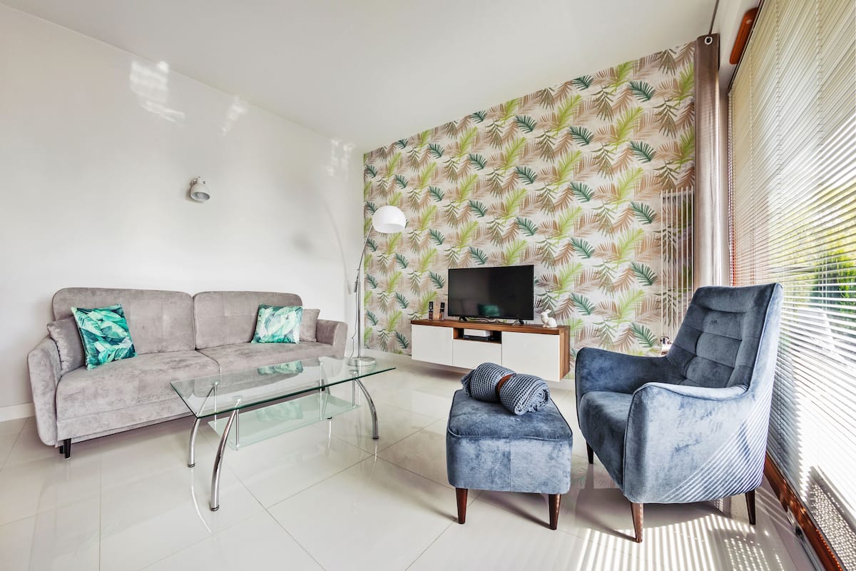 Un salon moderne avec un canapé gris, une table basse en verre, un fauteuil bleu et un téléviseur posé sur un meuble en bois.