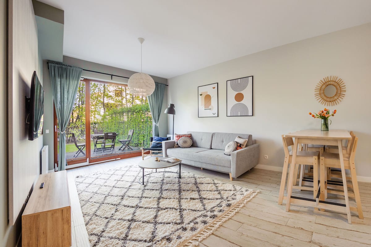 Un salon moderne avec un canapé, une table basse et un coin repas comprenant une table et des chaises en bois. Une grande fenêtre avec des rideaux, un luminaire au plafond et des œuvres d’art ornent le mur.