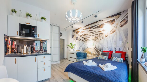 Habitación de hotel moderna con zona de cocina, cama con colcha azul y un diseño de techo único.