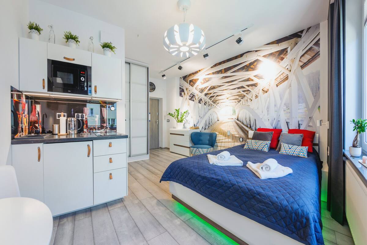A modern hotel room with a kitchen area, a bed with a blue quilt, and a unique ceiling design.