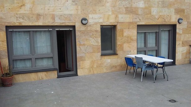 Un edificio con paredes de piedra, dos ventanas con cortinas, una mesa con sillas y una planta en maceta.