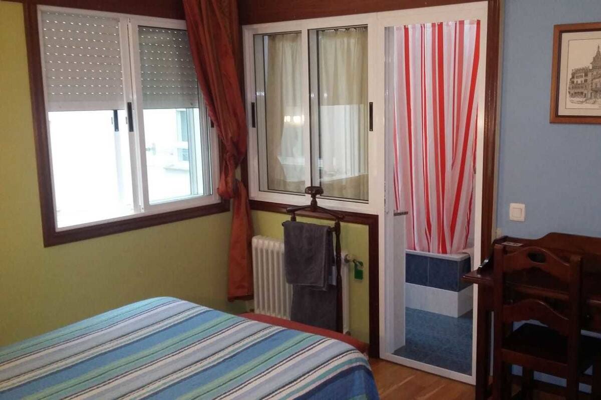 Quarto com cama, janela com cortinas, radiador, cadeira de madeira e um quadro emoldurado na parede.