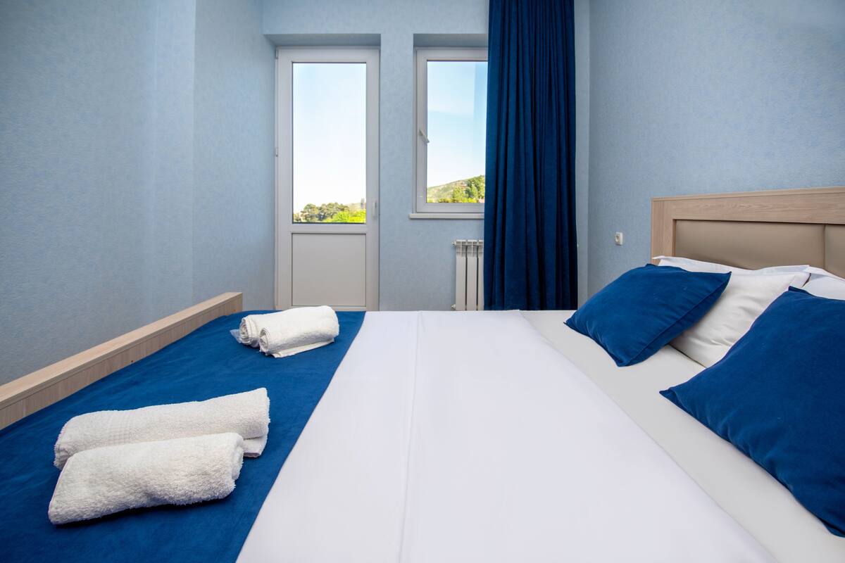 A hotel room with a large bed, two windows, a radiator, and a view of greenery outside.