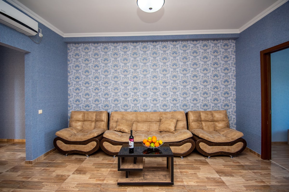 A living room with a brown sectional sofa, a black coffee table with a fruit bowl and a bottle, and patterned wallpaper.
