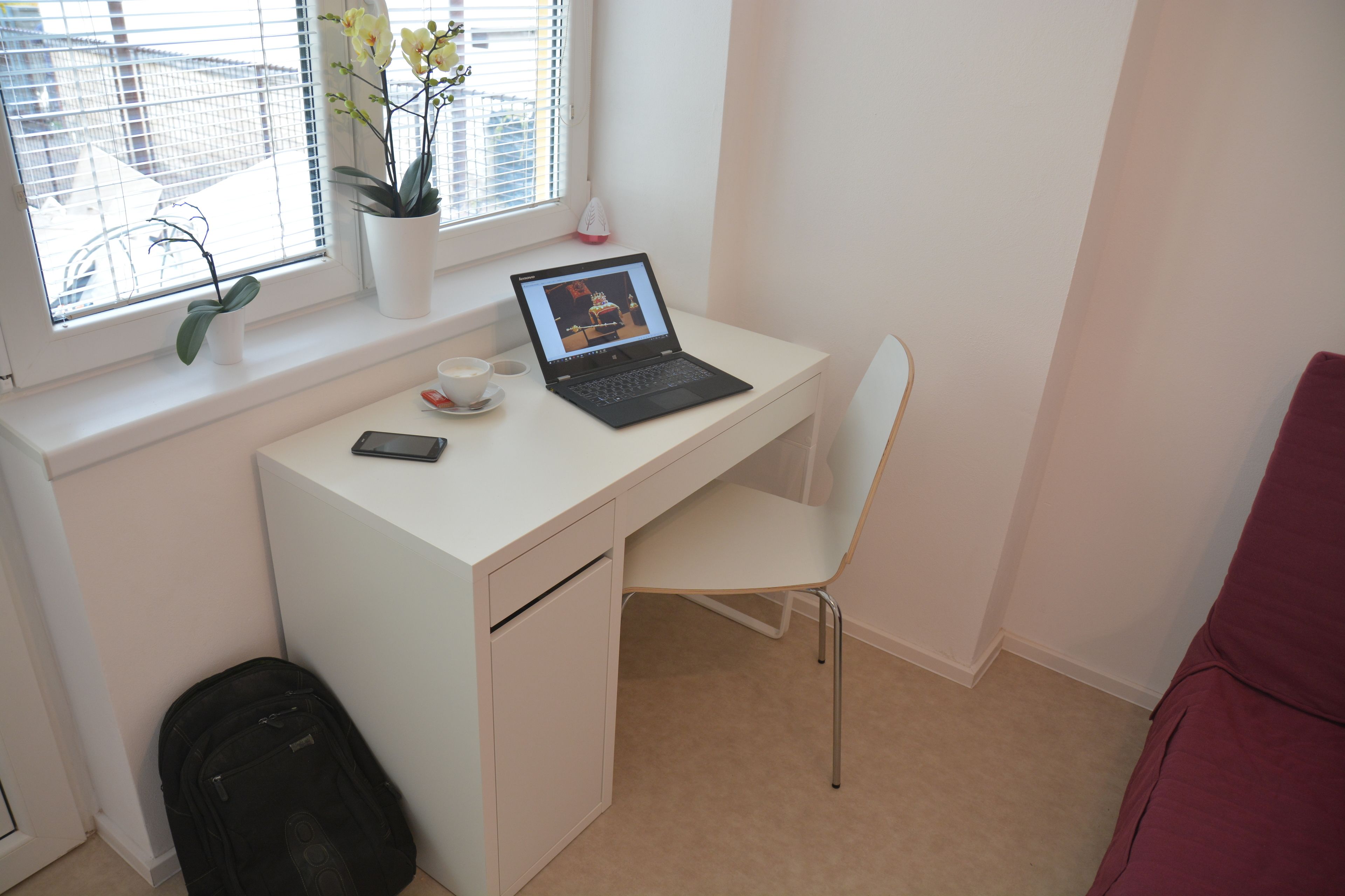Escritorio blanco con una computadora portátil, un teléfono inteligente y una taza. Se ve una ventana con plantas y una silla roja.