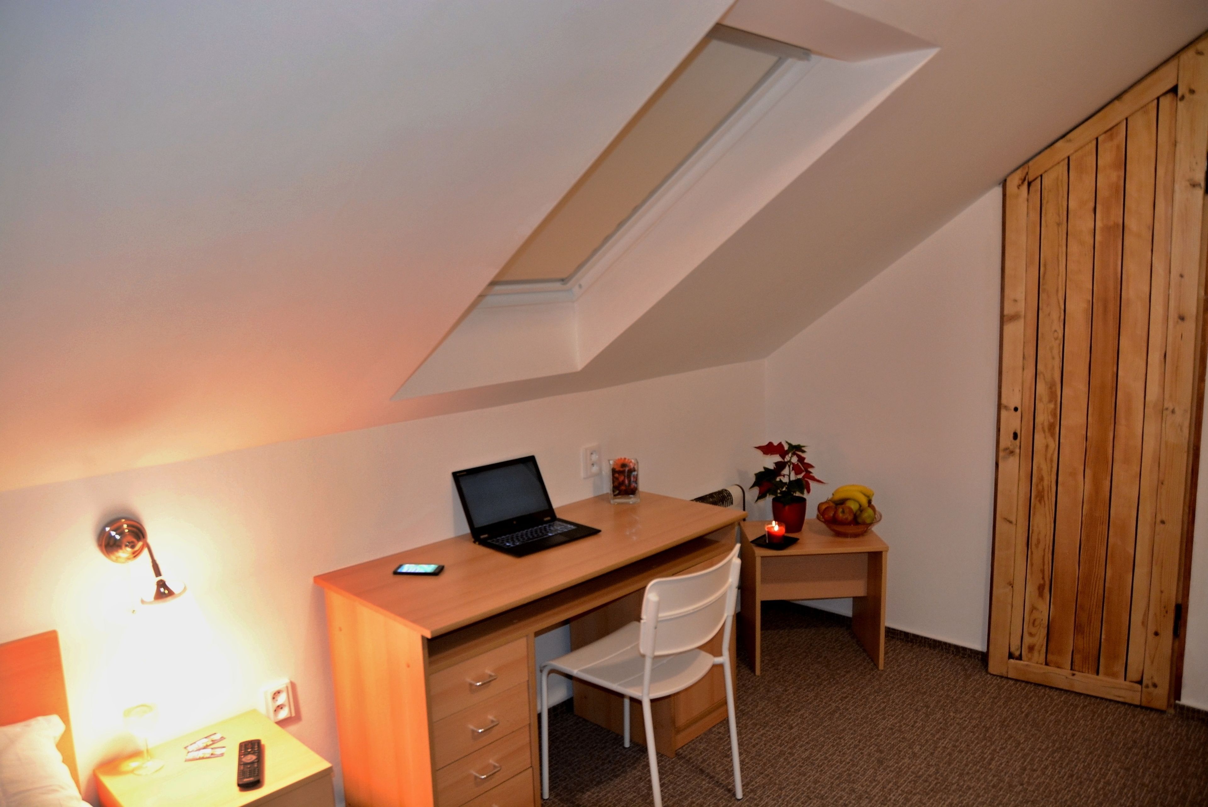 Quarto pequeno e bem iluminado, com uma escrivaninha de madeira, um laptop, uma cadeira branca, uma mesinha com uma fruteira e uma porta de madeira.