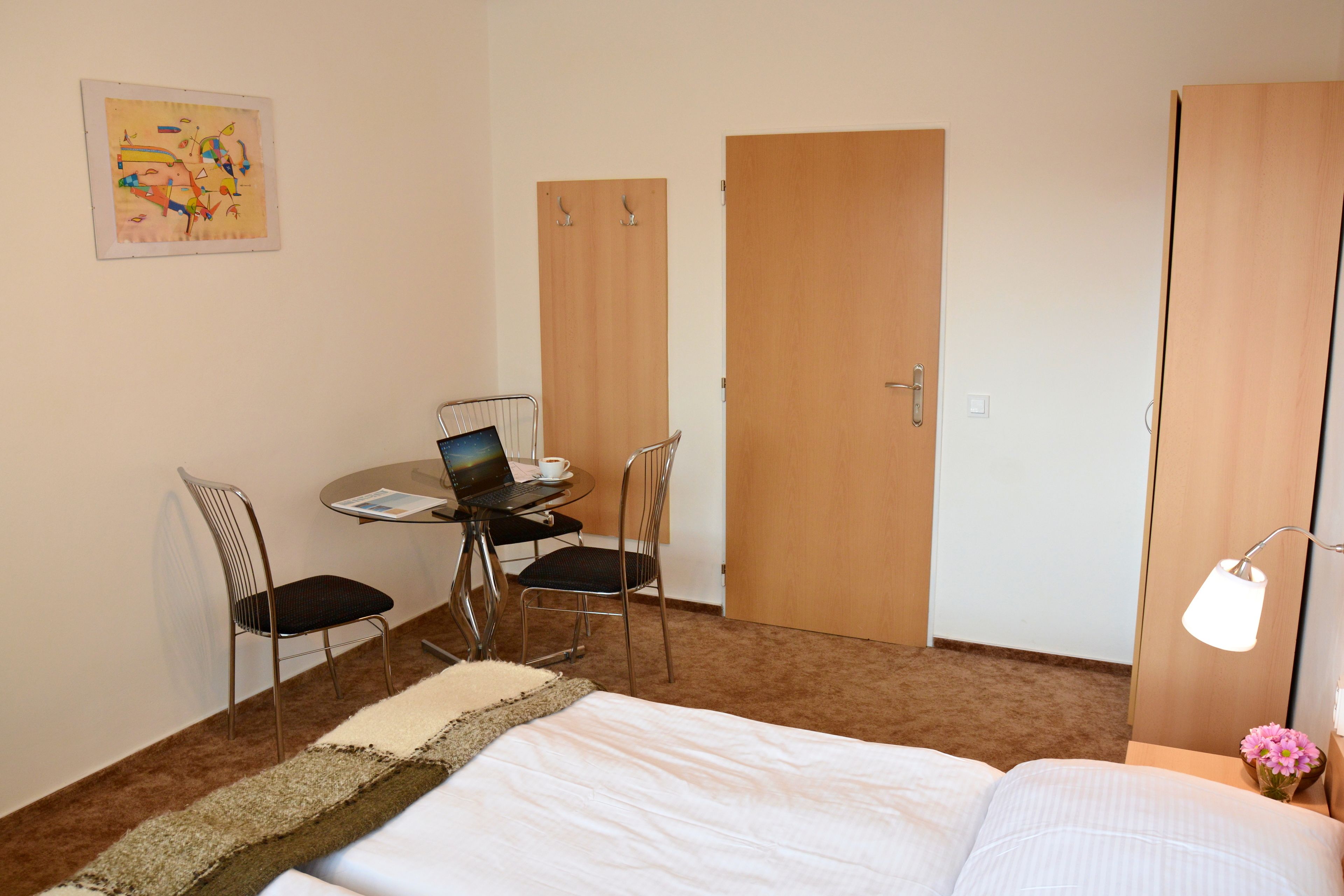 Una camera d'hotel con un letto, una scrivania con un portatile, due sedie, un tavolino con una tazza e una lampada.