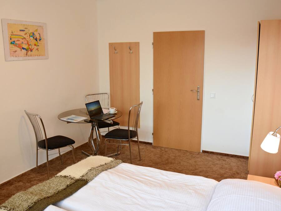 Una camera d'hotel con un letto, una scrivania con un portatile, due sedie, un tavolino con una tazza e una lampada.