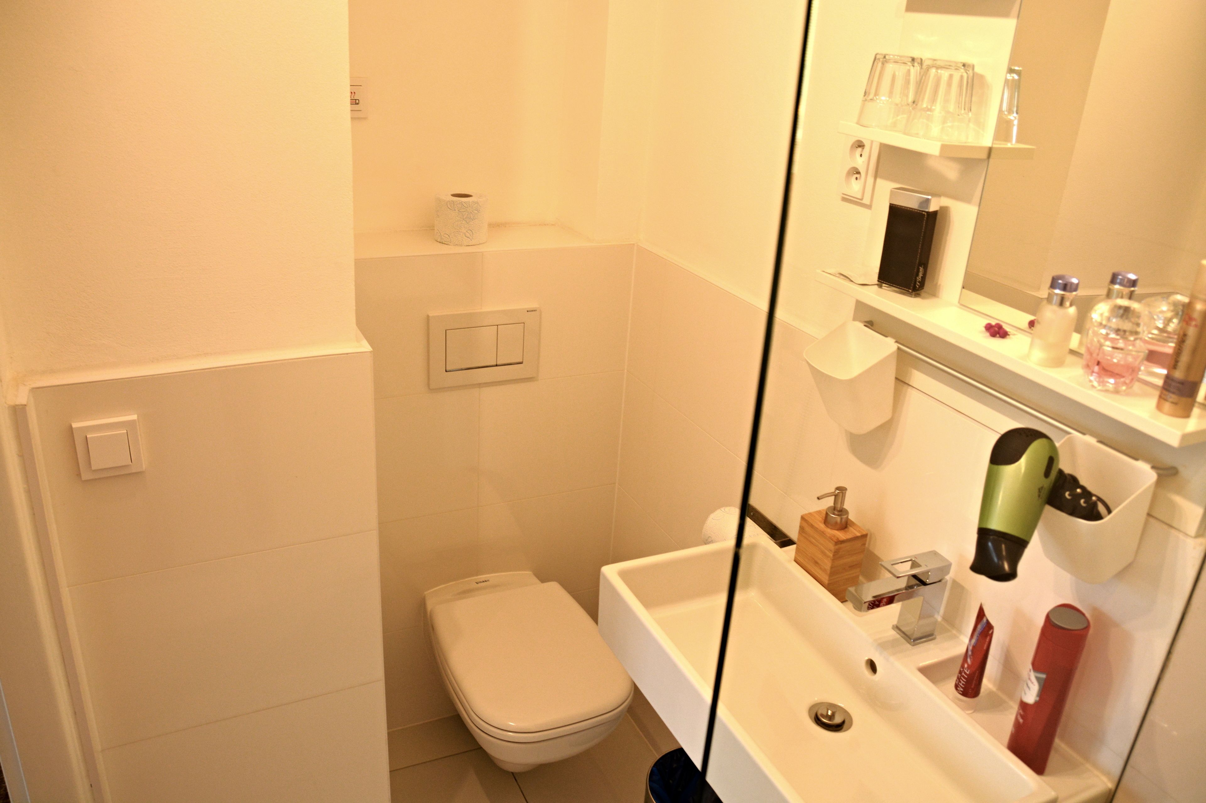 Ein kleines Badezimmer mit Toilette, Waschbecken und Dusche. Über dem Waschbecken befindet sich ein Spiegel und auf der Ablage sind Toilettenartikel zu sehen.