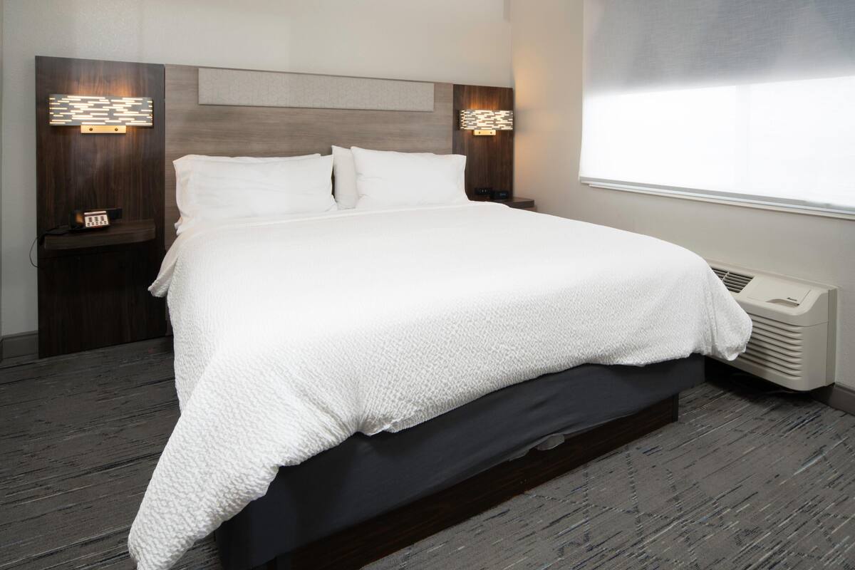 Una cama bien hecha con una manta blanca, un cabecero de madera, dos mesitas de noche con lámparas y una ventana con persianas.