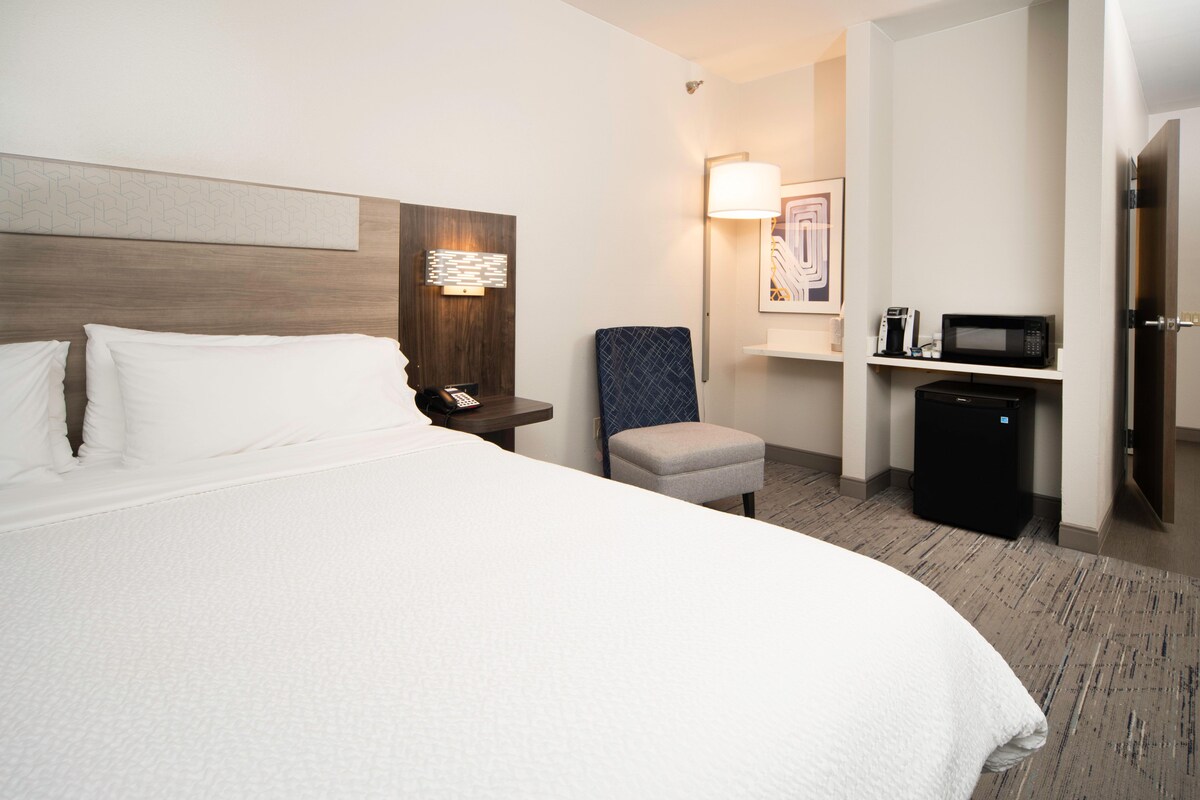 A hotel room with a large bed, a bedside table with a phone, a chair, a microwave, and a small refrigerator.