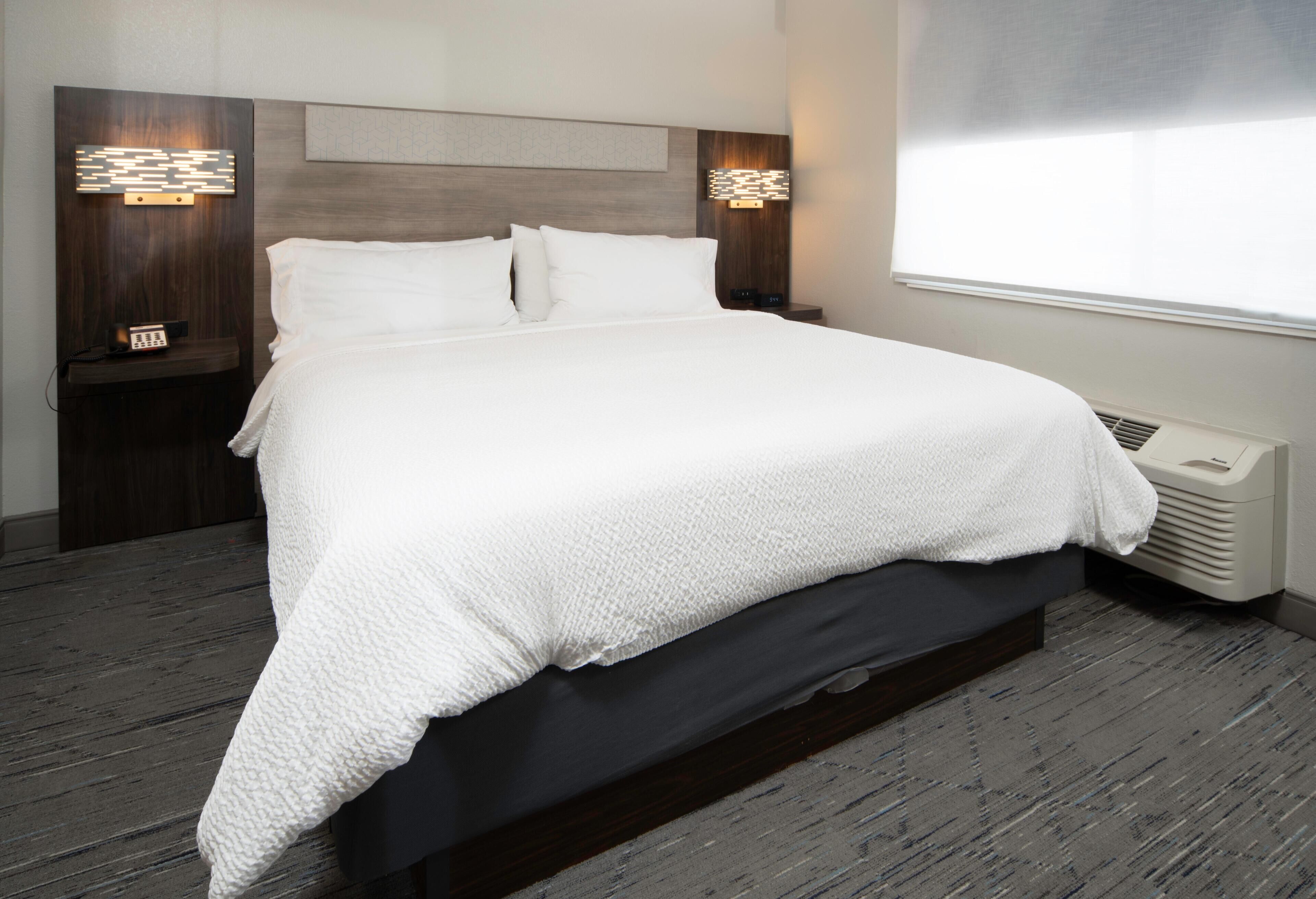 A neatly made bed with a white blanket, a wooden headboard, two bedside tables with lamps, and a window with blinds.