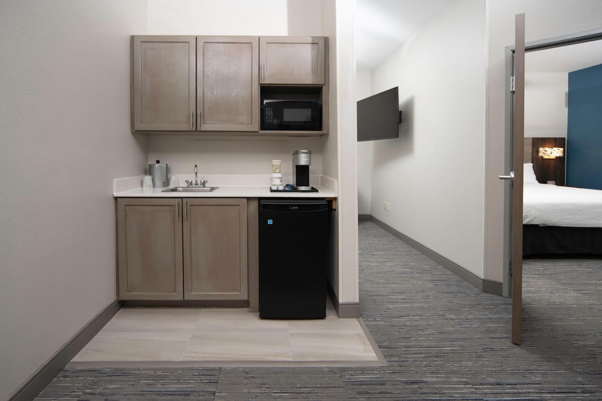 Una kitchenette compacta equipada con microondas, cafetera y lavavajillas, que da paso a un dormitorio con cama y televisor.