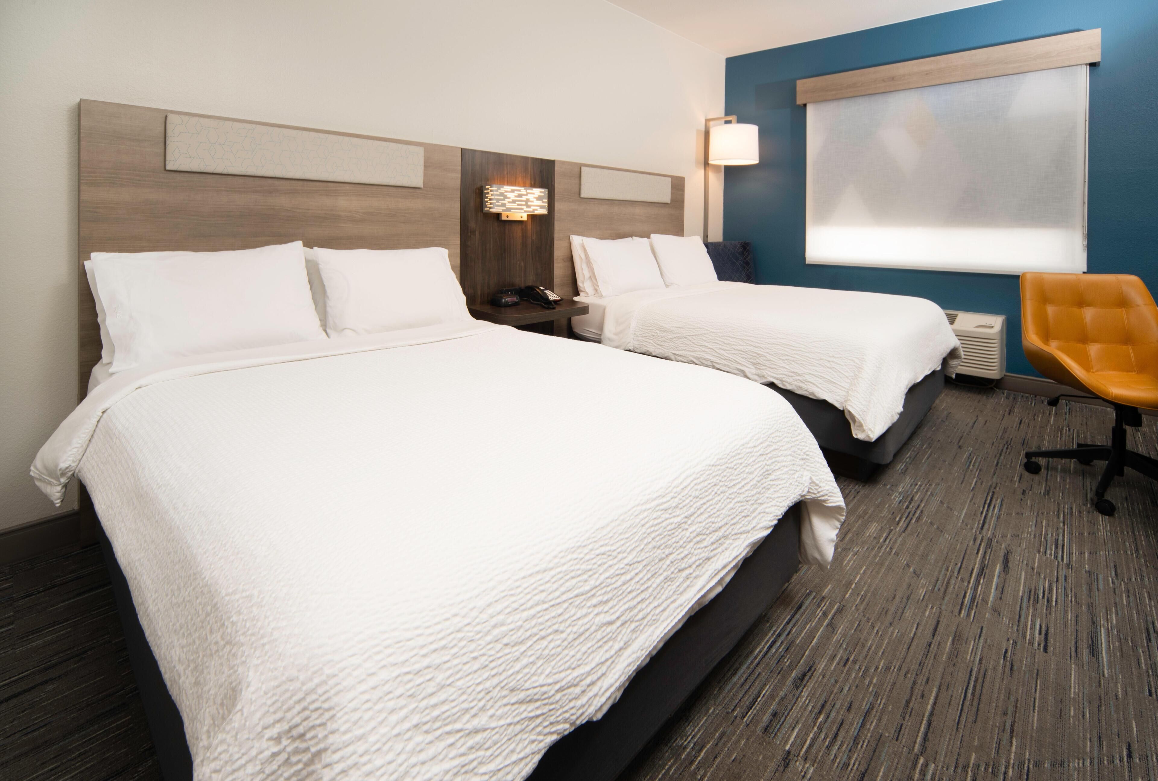 A hotel room with two beds, a nightstand, a lamp, a chair, and a window with blinds.
