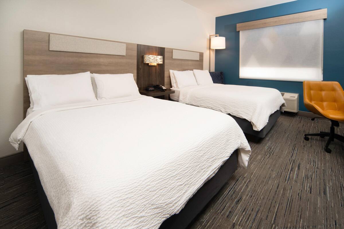 Una habitación de hotel con dos camas, una mesita de noche, una lámpara, una silla y una ventana con persianas.