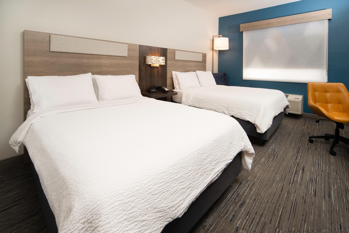 A hotel room with two beds, a nightstand, a lamp, a chair, and a window with blinds.
