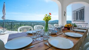 Ein Esstisch, gedeckt für sechs Personen, mit weißen Tellern, Gläsern und Besteck, eine Vase mit gelben Blumen und Blick auf die Landschaft.