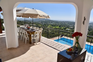 Une terrasse dotée d’une table, de chaises et d’un grand parasol, offrant une vue sur une piscine et un paysage pittoresque.